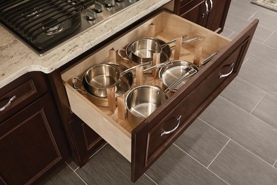 Pots and pans drawer base kitchen cabinet beneath a cooktop opened to show KraftMaid wood peg organizers containing pots and pans inside